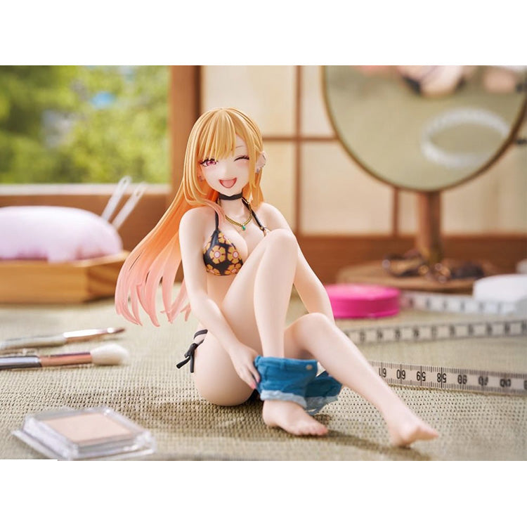Figure of Marin Kitagawa from My Dress-Up Darling sitting and smiling in a black floral swimsuit, holding blue shorts while posing playfully on a pink background.