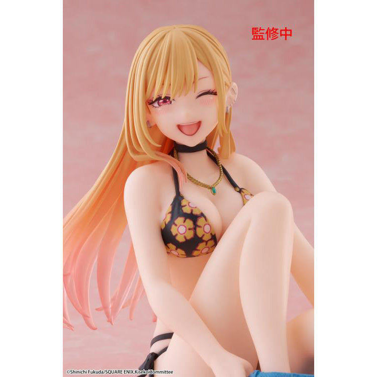 Figure of Marin Kitagawa from My Dress-Up Darling sitting and smiling in a black floral swimsuit, holding blue shorts while posing playfully on a pink background.